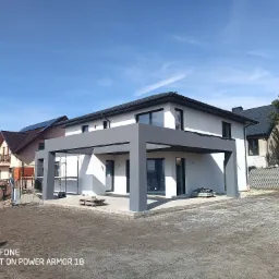 Nowoczesny dom jednorodzinny w Jaworznie z jasną elewacją i szarą pergolą w trakcie wykańczania, widoczne rusztowania i przygotowany teren.