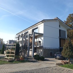 DANDI BUD Daniel Lejkowski - Dwupiętrowy dom w trakcie prac elewacyjnych, widoczne rusztowania i pracownicy na zewnątrz, częściowo otynkowana i pomalowana elewacja, teren wokół domu zagospodarowany kostką brukową i zielenią.