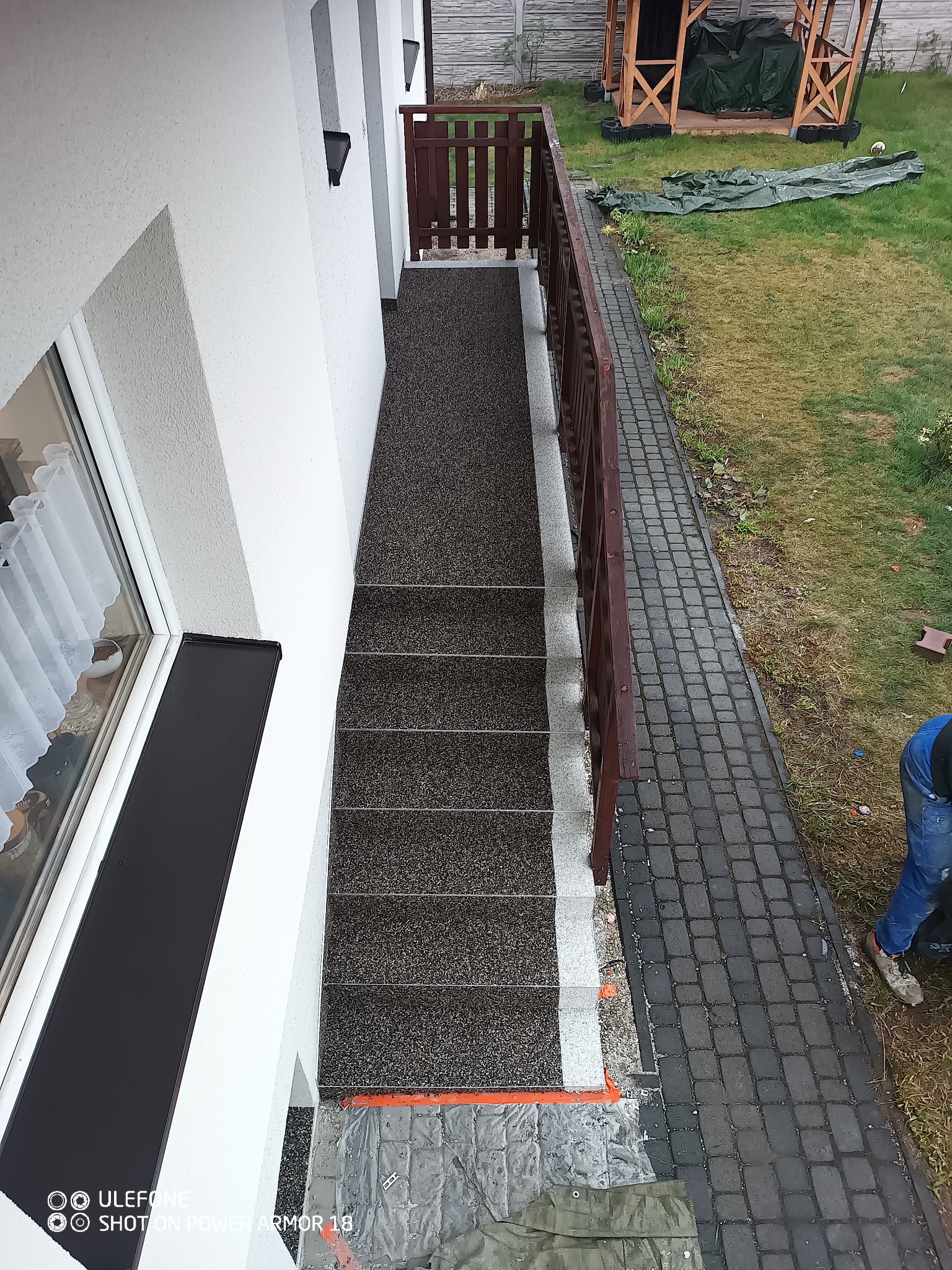 Zewnętrzne schody wejściowe pokryte nową, brązowo-szarą, żywiczną nawierzchnią z drobnym kruszywem, biała elewacja budynku, drewniana balustrada, widok z góry.