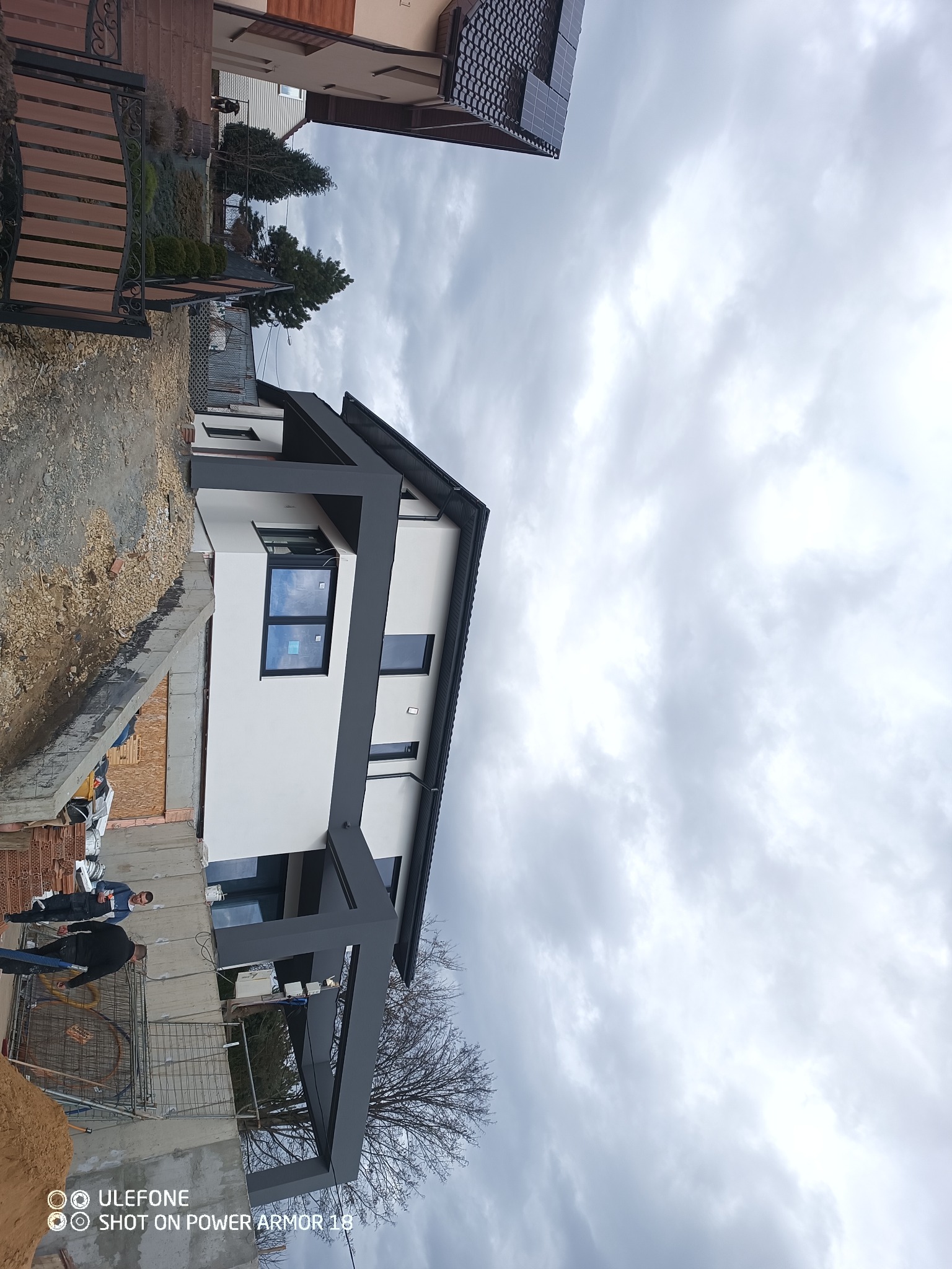 Nowoczesny dom jednorodzinny z białą elewacją i grafitowymi elementami wykończeniowymi, widoczny na etapie budowy z odsłoniętymi fundamentami i pracującymi osobami.