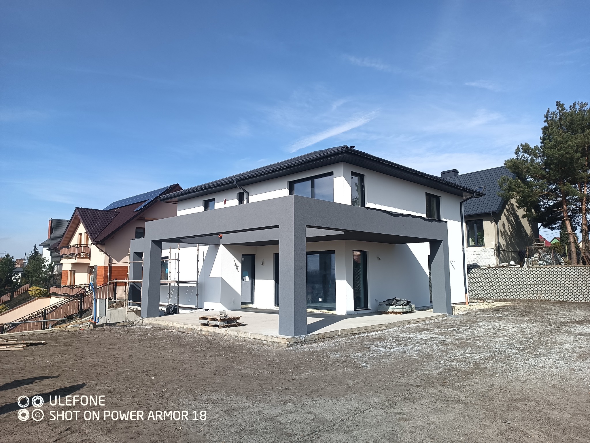 Nowoczesny dom jednorodzinny w Jaworznie z jasną elewacją i szarą pergolą w trakcie wykańczania, widoczne rusztowania i przygotowany teren.