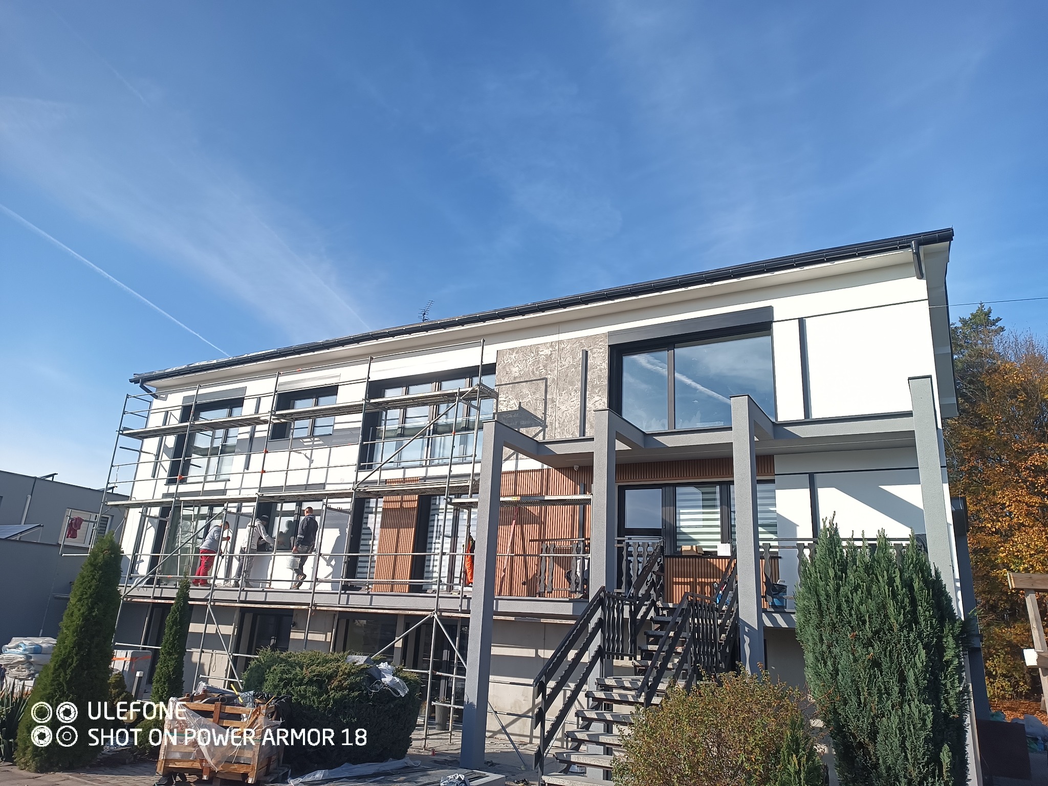 Dwukondygnacyjny dom z płaskim dachem w trakcie prac elewacyjnych, z widocznymi rusztowaniami, pracownikami i elementami dekoracyjnymi z drewnianymi panelami.