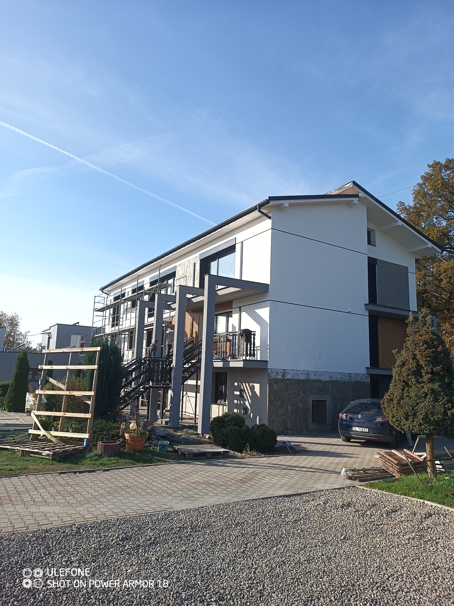 Dwupiętrowy dom w trakcie prac elewacyjnych, widoczne rusztowania i pracownicy na zewnątrz, częściowo otynkowana i pomalowana elewacja, teren wokół domu zagospodarowany kostką brukową i zielenią.