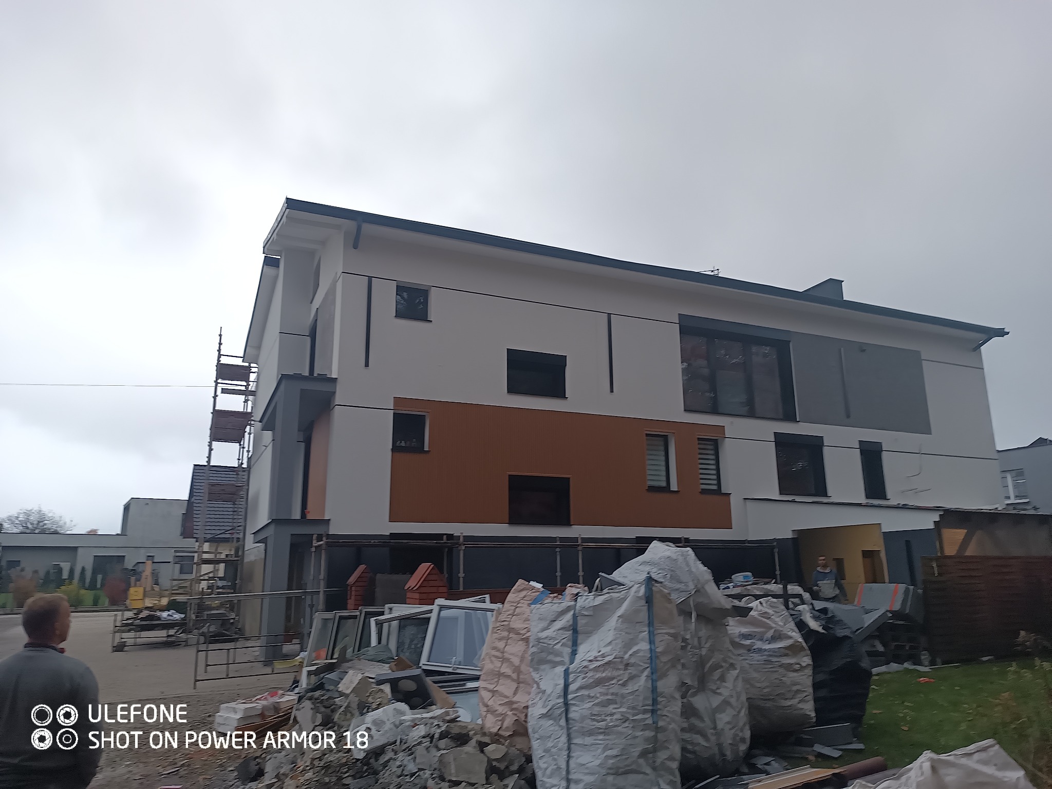 Dwukondygnacyjny dom z częściowo wykonaną elewacją w kolorach bieli i brązu, widoczne rusztowania, okna i worki z gruzem na pierwszym planie.