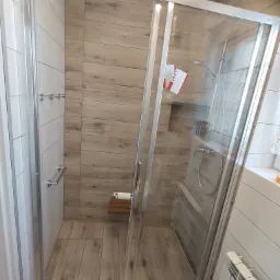Kabina prysznicowa z przesuwnymi drzwiami, drewnopodobne płytki na ścianach i podłodze, małe składane siedzisko wewnątrz kabiny, widoczny grzejnik na ścianie obok.