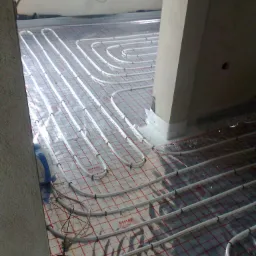 Białe rury ogrzewania podłogowego ułożone na folii z siatką, widoczne elementy mocujące i podłączone przewody, w tle fragment ściany i słupa.