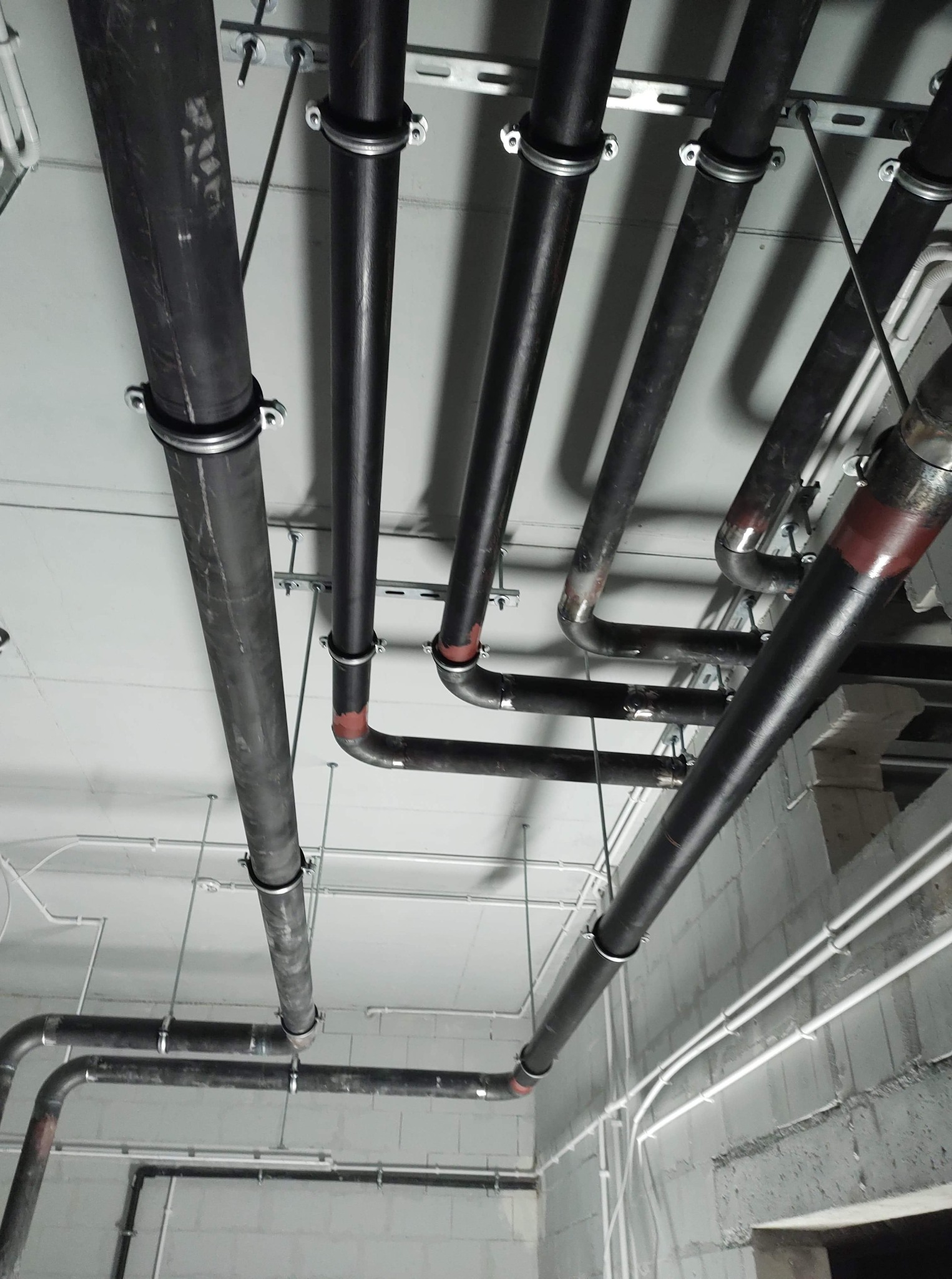 Skomplikowany system czarnych rur grzewczych i hydraulicznych pod sufitem z widocznymi mocowaniami, niektóre rury z czerwonymi oznaczeniami, instalacja w surowym stanie.