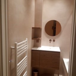 Heli-Bet - Nowoczesna łazienka z beżowymi ścianami, okrągłym lustrem, umywalką z szafką z drewnianym fornirem, grzejnikiem drabinkowym i koszem na śmieci w kolorze miedzi.