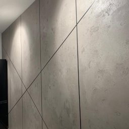 Heli-Bet - Ściana z szarego betonu architektonicznego z czarnymi, ukośnymi listwami dekoracyjnymi podświetlona punktowymi lampami sufitowymi.