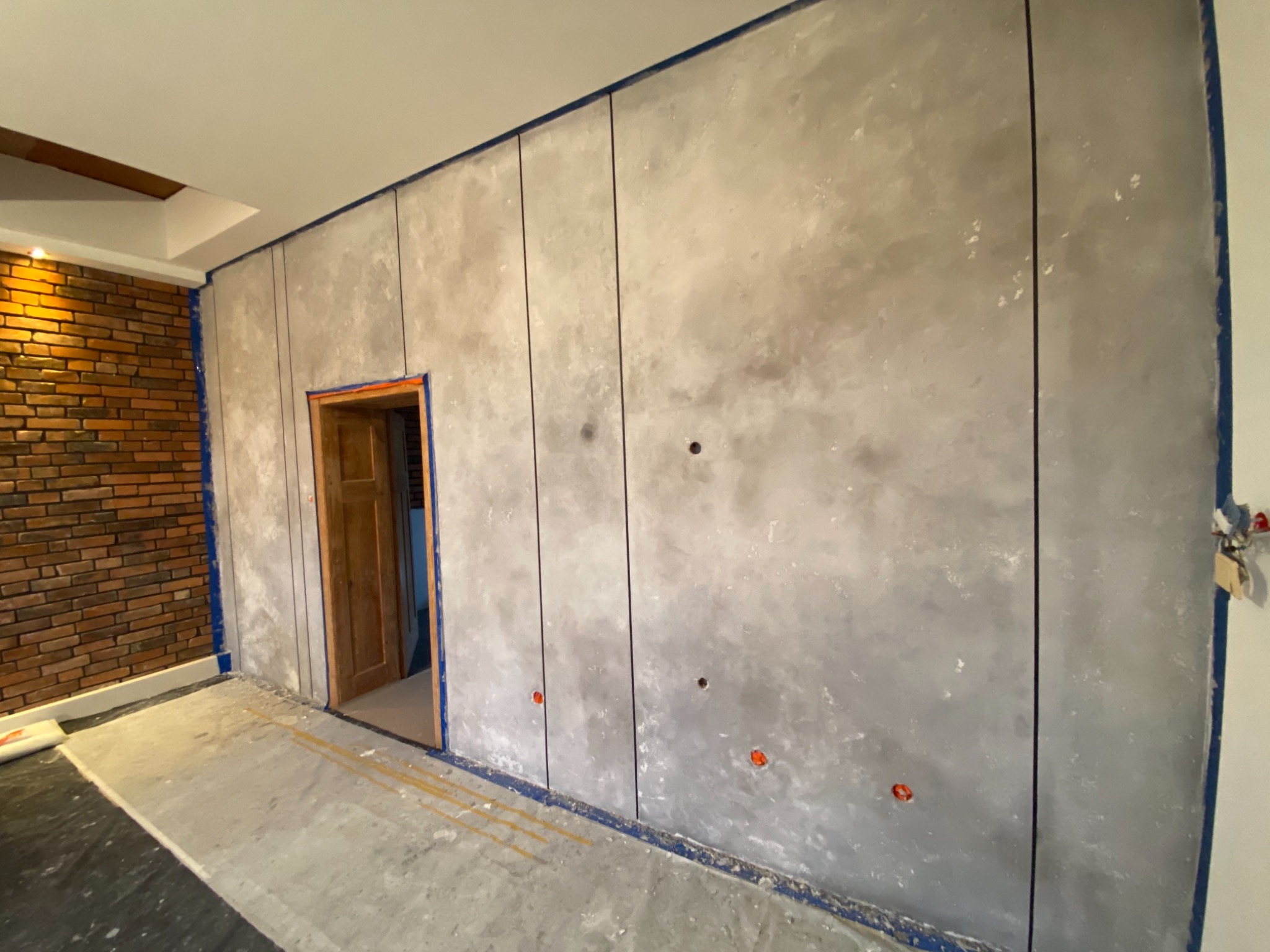 Ściana z dekoracyjnym tynkiem imitującym beton, podzielona pionowymi liniami, zabezpieczona niebieską taśmą malarską, widoczne otwory na gniazdka elektryczne, fragment ściany z cegły w tle.