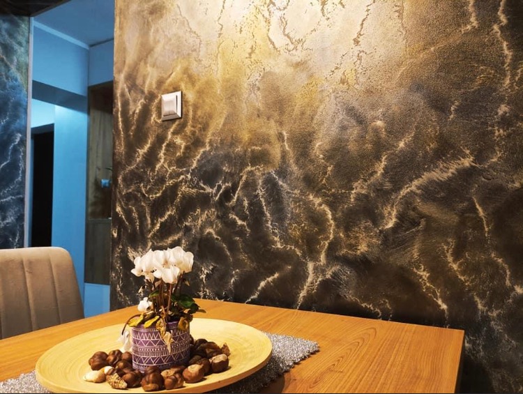 Dekoracyjna ściana z efektem marmuru w odcieniach brązu i złota, oświetlona z góry, z włącznikiem światła, na stole drewniana misa z kasztanami i kwiatami.