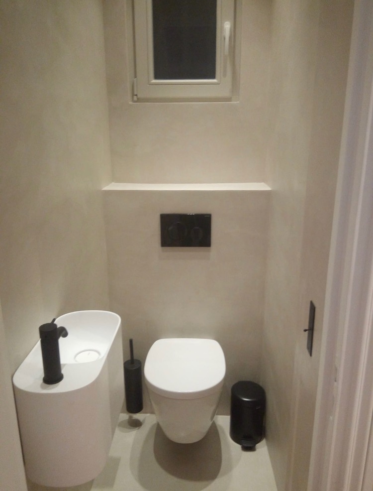 Nowoczesna, minimalistyczna toaleta z podwieszaną miską WC i umywalką nablatową o organicznym kształcie, czarna armatura, beżowe ściany z fakturą, małe okno w górnej części ściany.