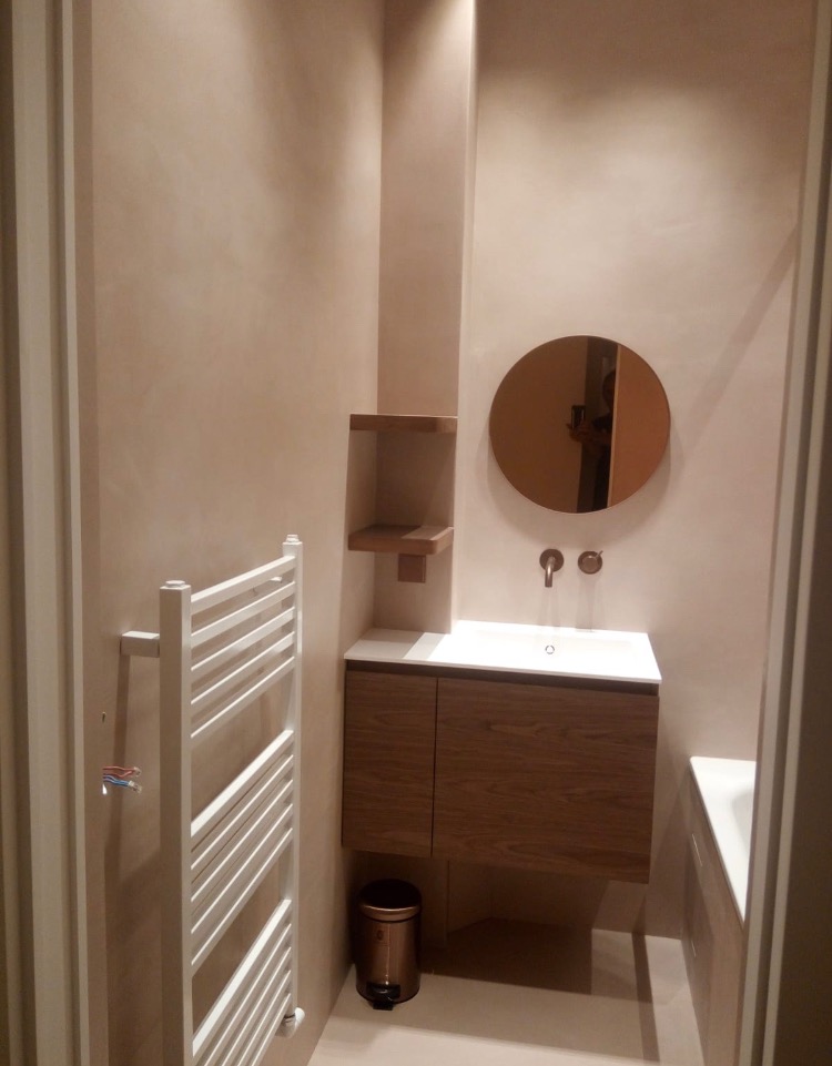 Nowoczesna łazienka z beżowymi ścianami, okrągłym lustrem, umywalką z szafką z drewnianym fornirem, grzejnikiem drabinkowym i koszem na śmieci w kolorze miedzi.