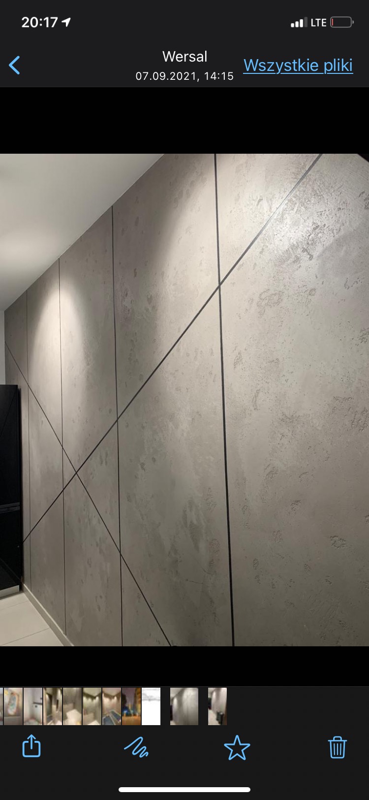 Ściana z szarego betonu architektonicznego z czarnymi, ukośnymi listwami dekoracyjnymi podświetlona punktowymi lampami sufitowymi.