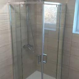 Nowoczesna kabina prysznicowa z chromowaną armaturą, jasnymi płytkami imitującymi drewno i widokiem na drzewa za oknem.