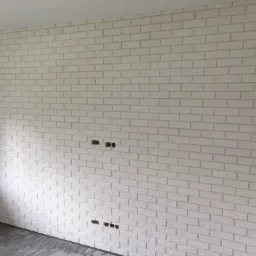 Ściana wyłożona białymi, cegiełkami z widocznymi otworami na gniazdka elektryczne, podłoga częściowo pokryta szarą wylewką.