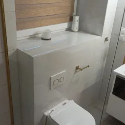 Nowoczesna toaleta podwieszana z zabudową geberit obłożoną płytkami imitującymi marmur, z akcentami w kolorze złotym: przycisk spłuczki, uchwyt na papier toaletowy i rama kabiny prysznicowej.