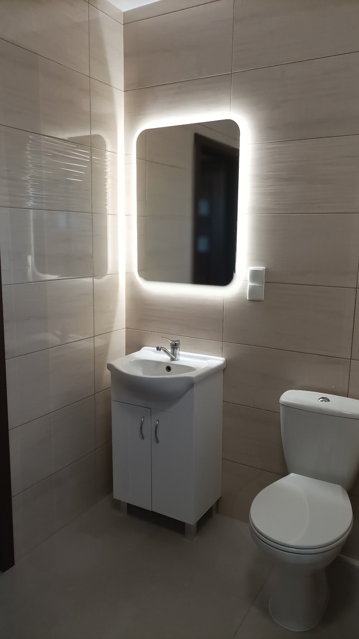 Ukończona łazienka z beżowymi płytkami, podświetlanym lustrem nad białą szafką z umywalką i toaletą.