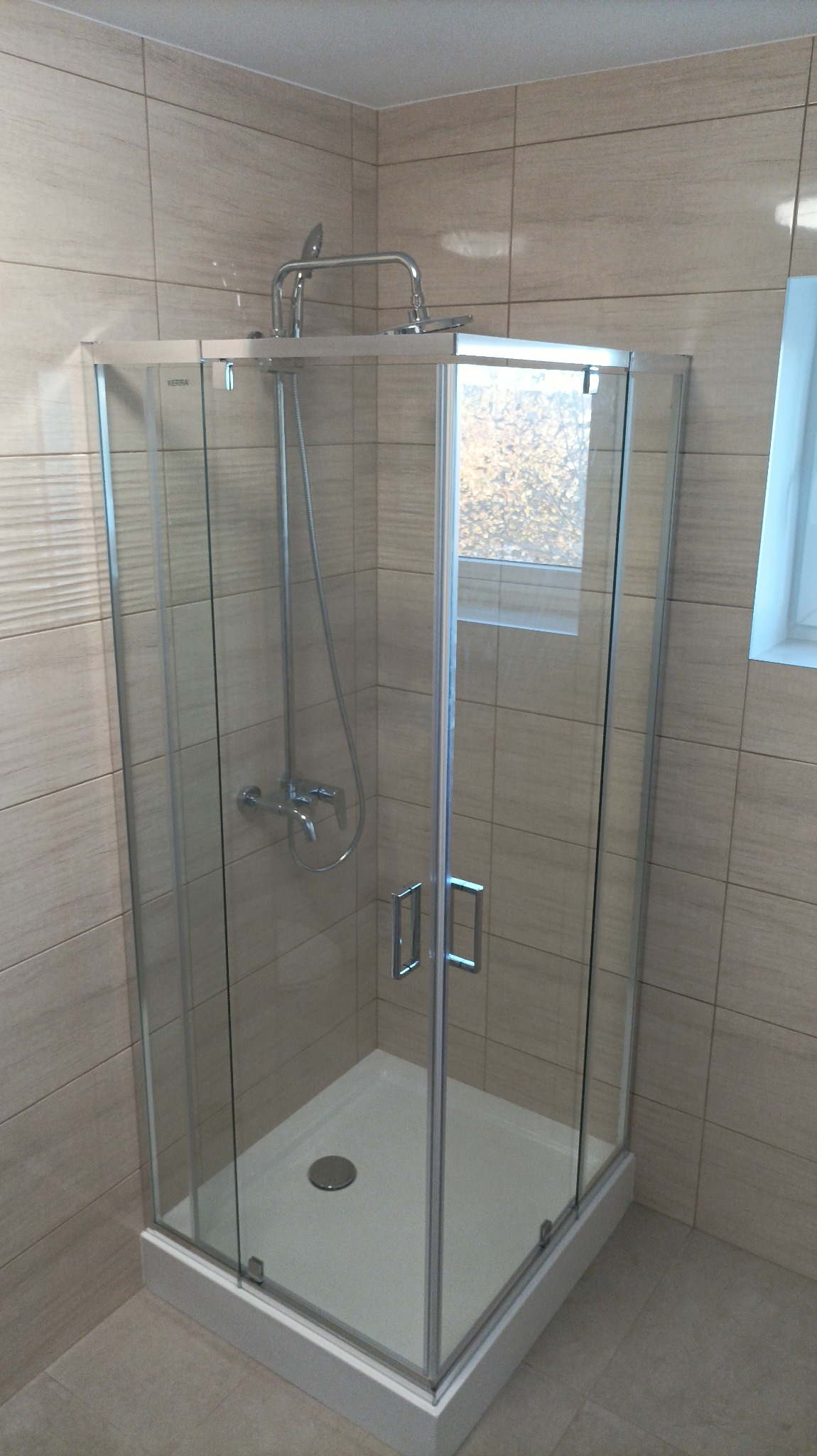 Nowoczesna kabina prysznicowa z chromowaną armaturą, jasnymi płytkami imitującymi drewno i widokiem na drzewa za oknem.