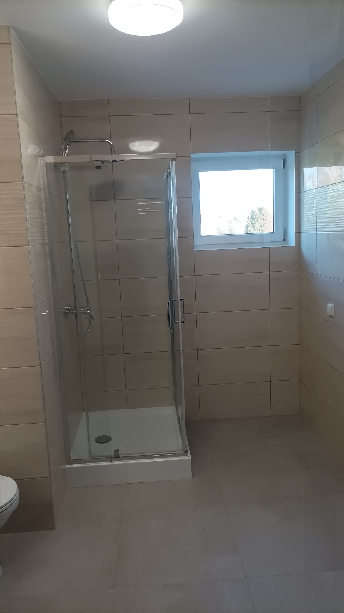 Nowoczesna kabina prysznicowa z transparentnego szkła, jasne płytki ceramiczne na ścianach i podłodze, okno z widokiem na drzewa, minimalistyczne oświetlenie sufitowe.