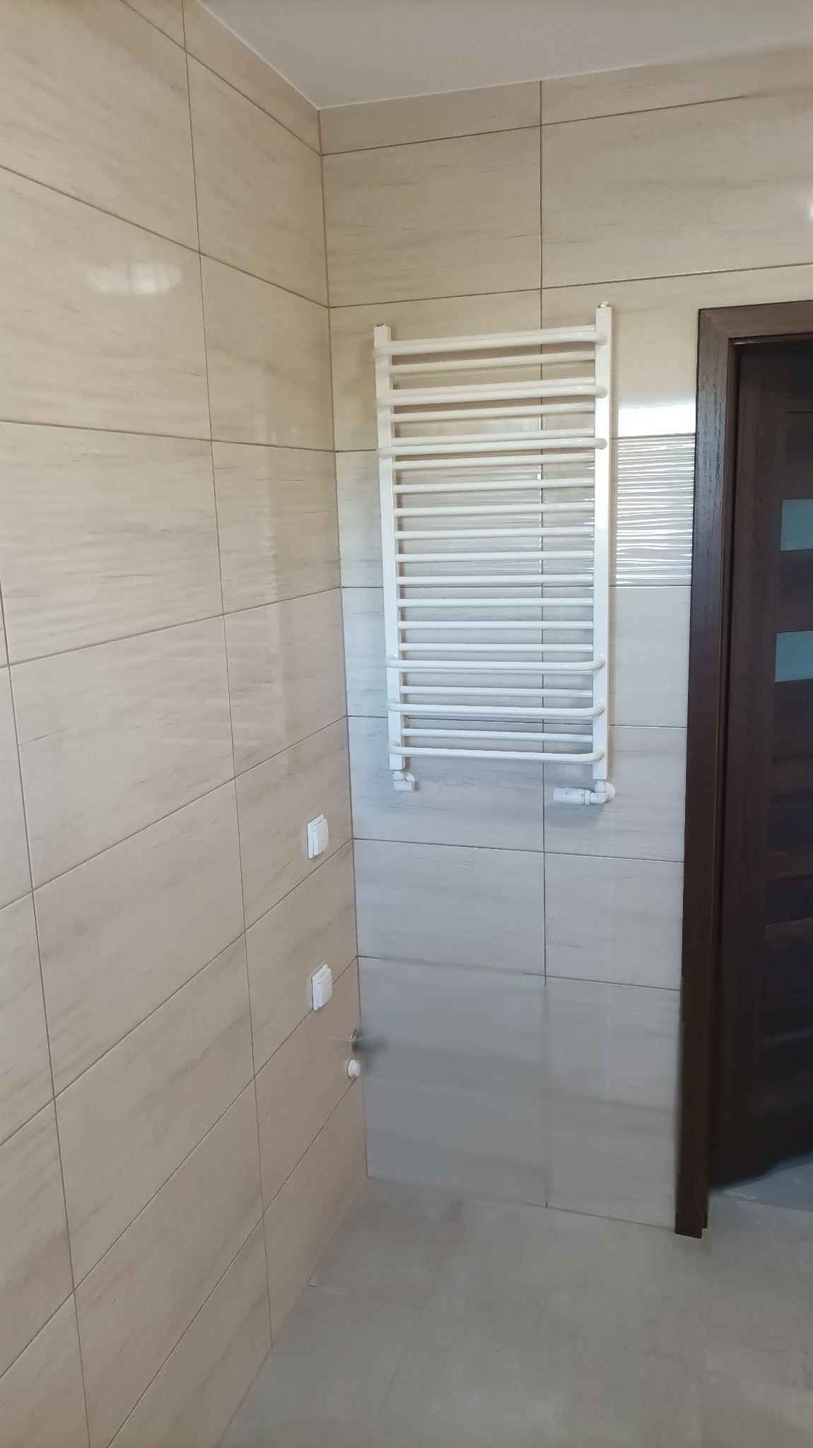 Biały grzejnik drabinkowy zamontowany na beżowych, poziomych płytkach w łazience, widoczny fragment ciemnobrązowych drzwi.