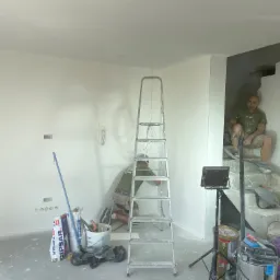 Dwóch mężczyzn pracuje w jasnym pomieszczeniu: jeden szpachluje ścianę przy drabinie, drugi siedzi na betonowych schodach w szarej koszulce, w tle worki z gipsem i narzędzia.