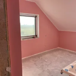 Poddasze po malowaniu na różowo, widoczne niedoskonałości podłogi i fragment niezabezpieczonej ściany z cegły.