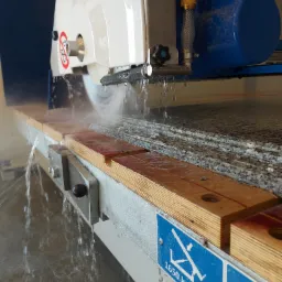 Cięcie kamienia na mokro za pomocą piły diamentowej; woda chłodzi ostrze i usuwa urobek, płyty kamienne ułożone warstwowo na stole roboczym, widoczna tabliczka informacyjna o maksymalnym obciążeniu.