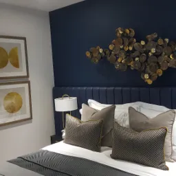 Nowoczesna sypialnia z dekoracyjną ścianą w kolorze granatowym, ozdobiona abstrakcyjną metalową kompozycją nad wezgłowiem łóżka, uzupełniona o minimalistyczne obrazy w złotych ramach na białej...