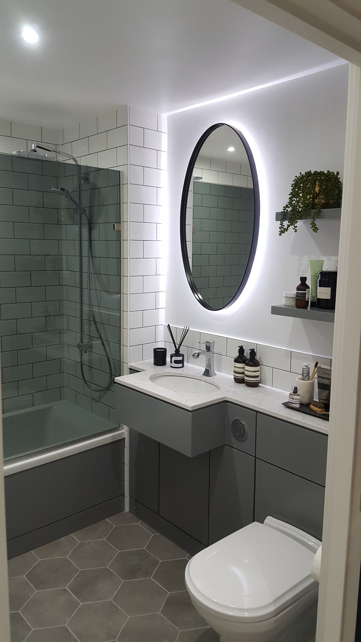 Nowoczesna łazienka w szarościach z heksagonalną podłogą, podświetlanym owalnym lustrem, białą armaturą i minimalistycznymi półkami z dekoracjami.