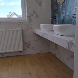 Bud-Mar Marcin Brzeziński - Nowoczesna łazienka z drewnopodobną podłogą, białymi umywalkami nablatowymi na marmurowym blacie, płytkami imitującymi marmur na ścianach i dekoracyjną ścianą z motywem liści oraz okrągłym lustrem.