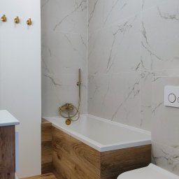 Bud-Mar Marcin Brzeziński - Nowoczesna łazienka z wanną obudowaną drewnopodobnymi panelami, marmurowe płytki na ścianach i złota armatura, tworzą elegancki i spójny wygląd.