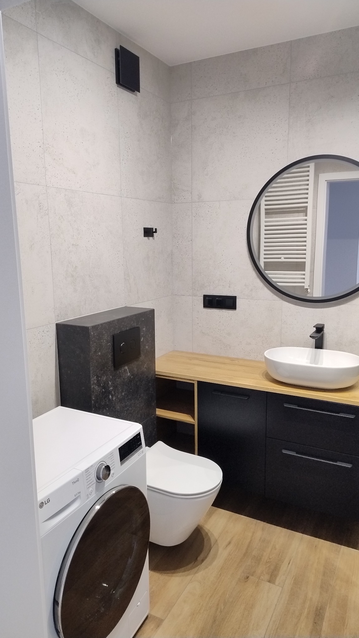 Nowoczesna łazienka z beżowymi płytkami, podwieszaną toaletą z czarnym panelem, okrągłym lustrem i pralką LG, drewnopodobna podłoga.