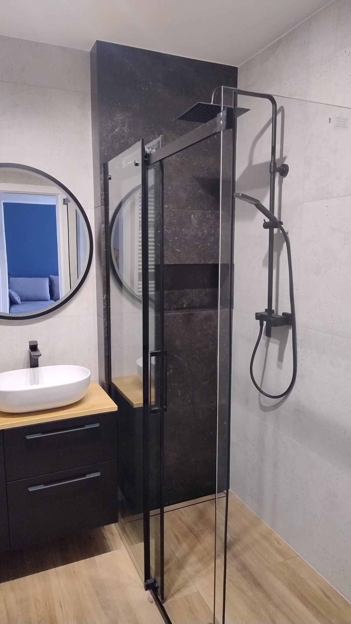 Nowoczesna łazienka z kabiną prysznicową typu walk-in z czarnymi profilami, owalną umywalką nablatową i okrągłym lustrem, widok przez otwarte drzwi na pokój z szarą sofą.