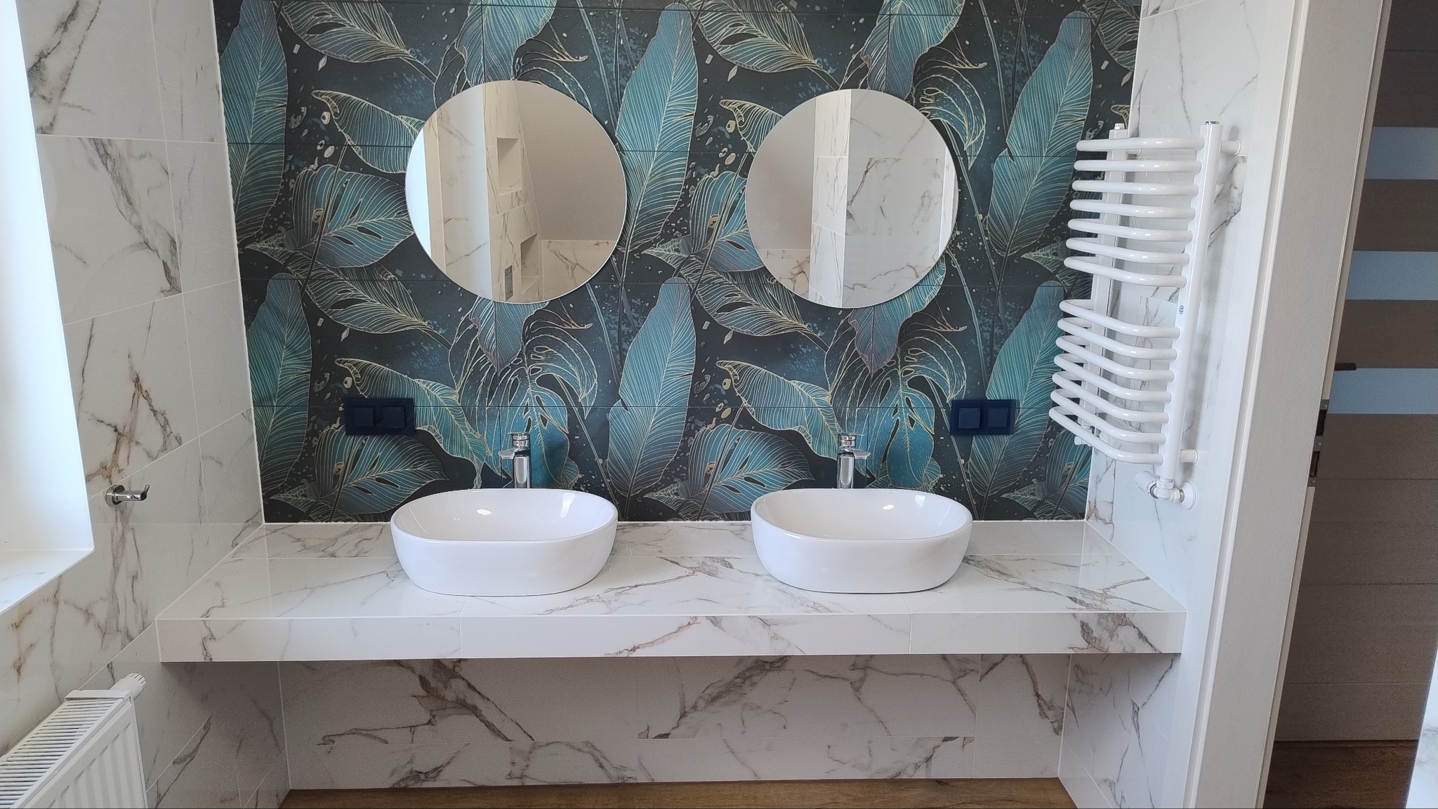 Nowoczesna łazienka z dwiema owalnymi umywalkami nablatowymi, okrągłymi lustrami i dekoracyjną tapetą w motywy roślinne, uzupełniona grzejnikiem drabinkowym.