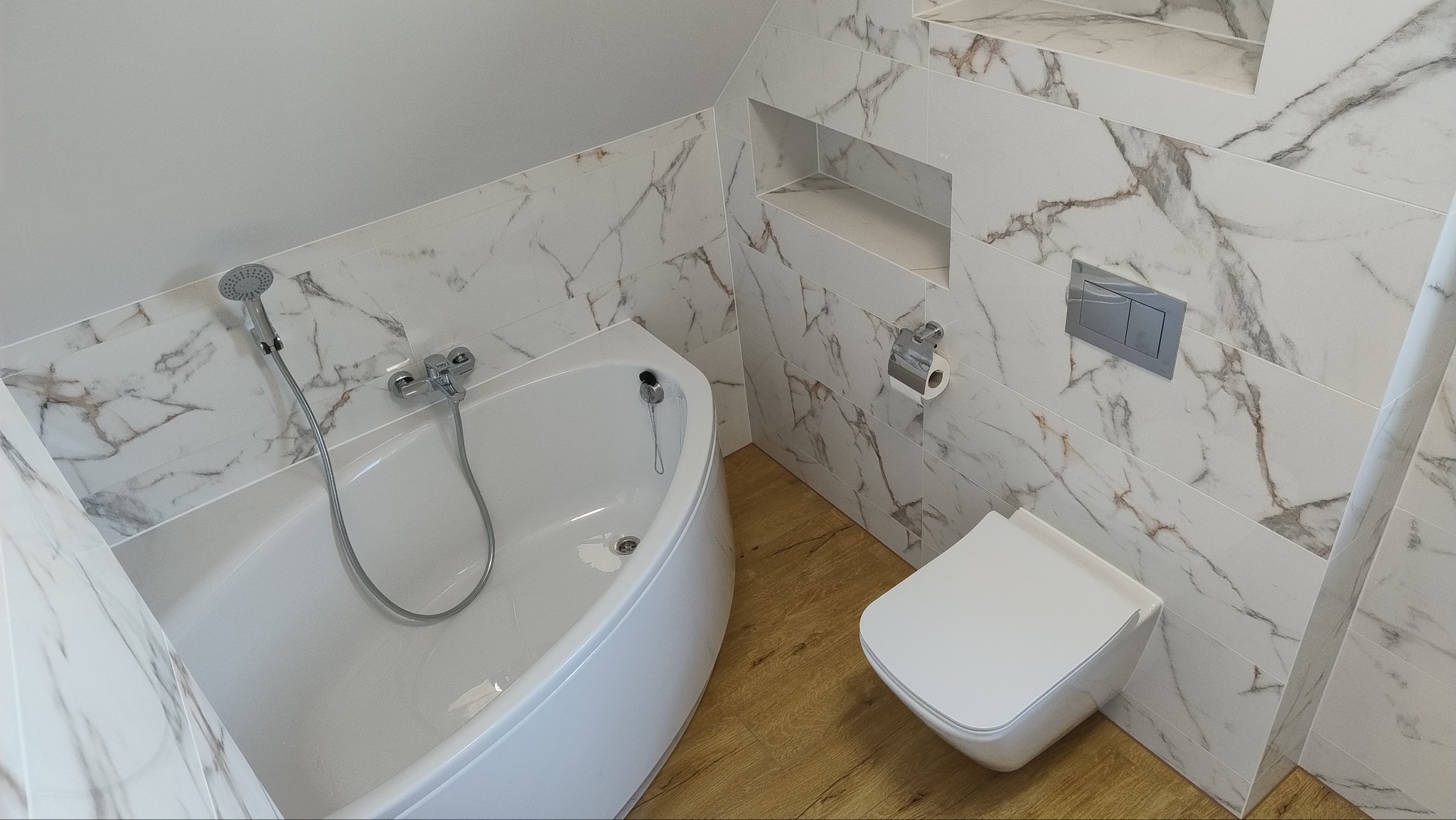 Nowoczesna łazienka z wanną narożną, toaletą podwieszaną i płytkami imitującymi marmur, drewnopodobna podłoga, wnęka w ścianie.