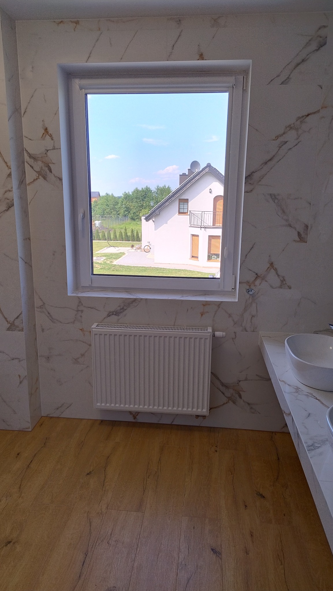 Łazienka z marmurowymi płytkami na ścianach, drewnopodobną podłogą, białym grzejnikiem pod oknem z widokiem na dom i błękitne niebo.