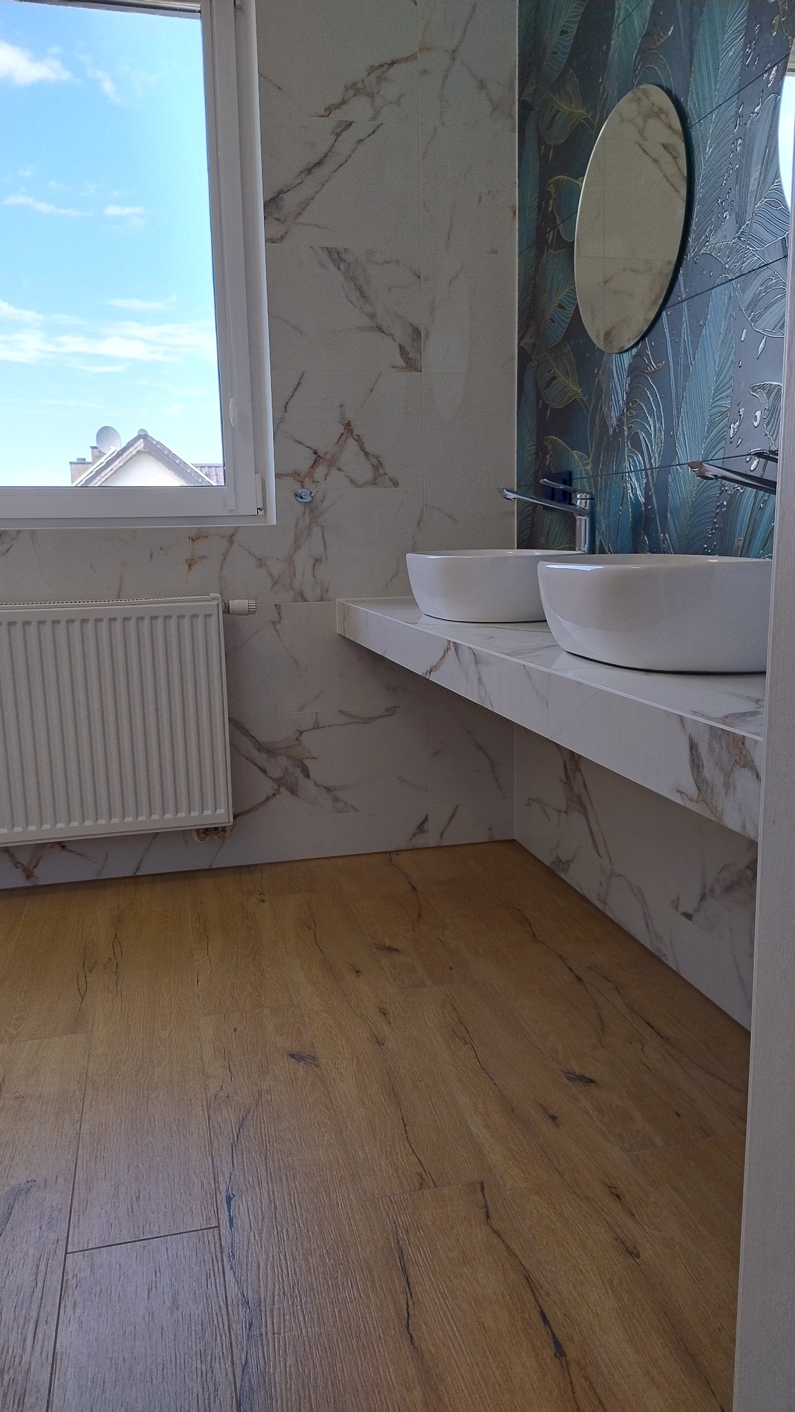 Nowoczesna łazienka z drewnopodobną podłogą, białymi umywalkami nablatowymi na marmurowym blacie, płytkami imitującymi marmur na ścianach i dekoracyjną ścianą z motywem liści oraz okrągłym lustrem.