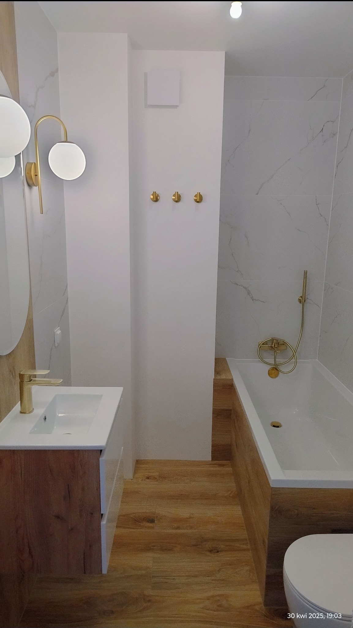 Nowoczesna łazienka z drewnopodobną podłogą, białą armaturą i złotymi akcentami: bateria umywalkowa, słuchawka prysznicowa, wieszaki. Minimalistyczny design.