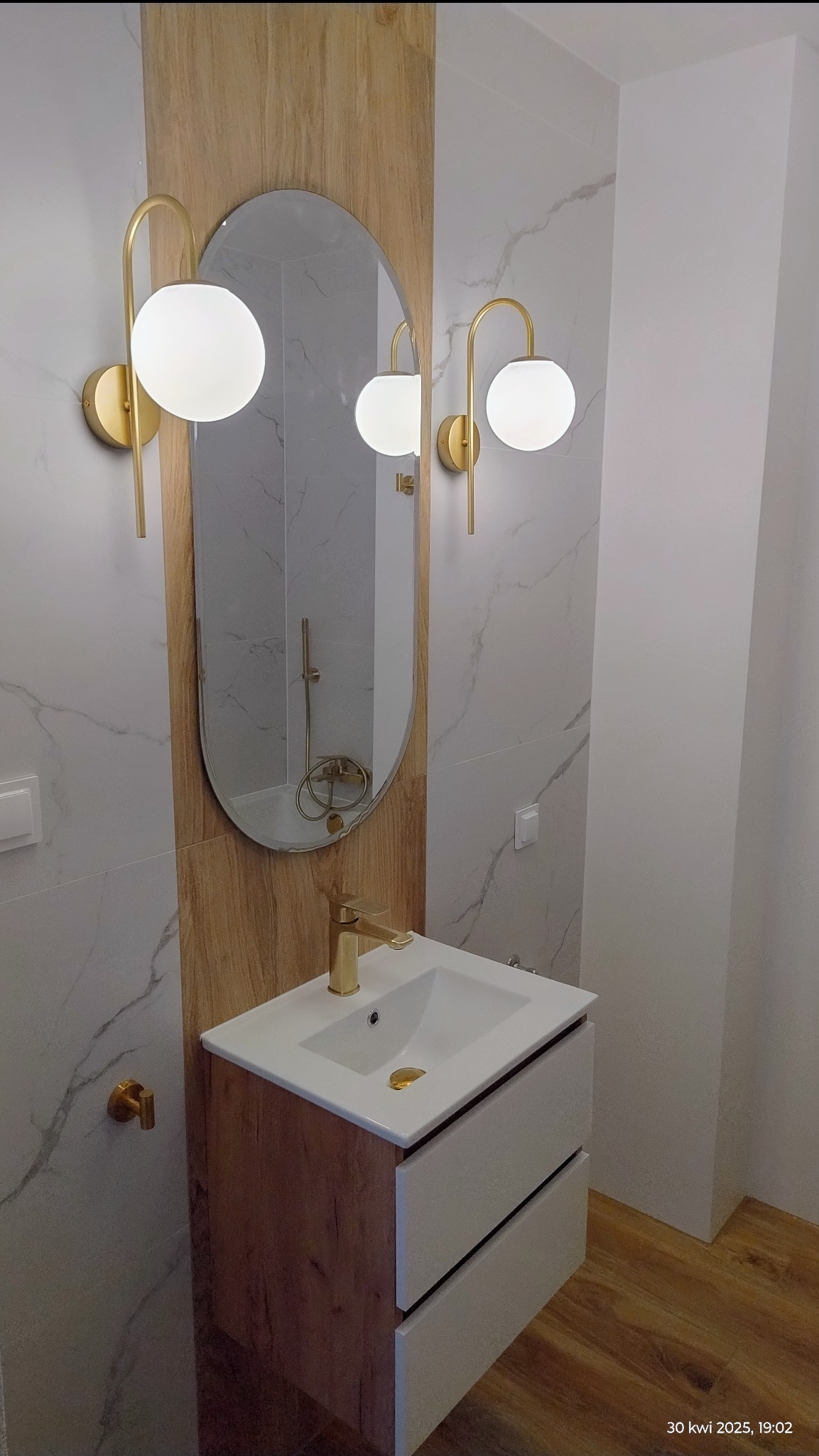 Nowoczesna łazienka: biała umywalka z szafką w odcieniach drewna, złota armatura i owalne lustro podświetlone kulistymi lampami na marmurowej ścianie.