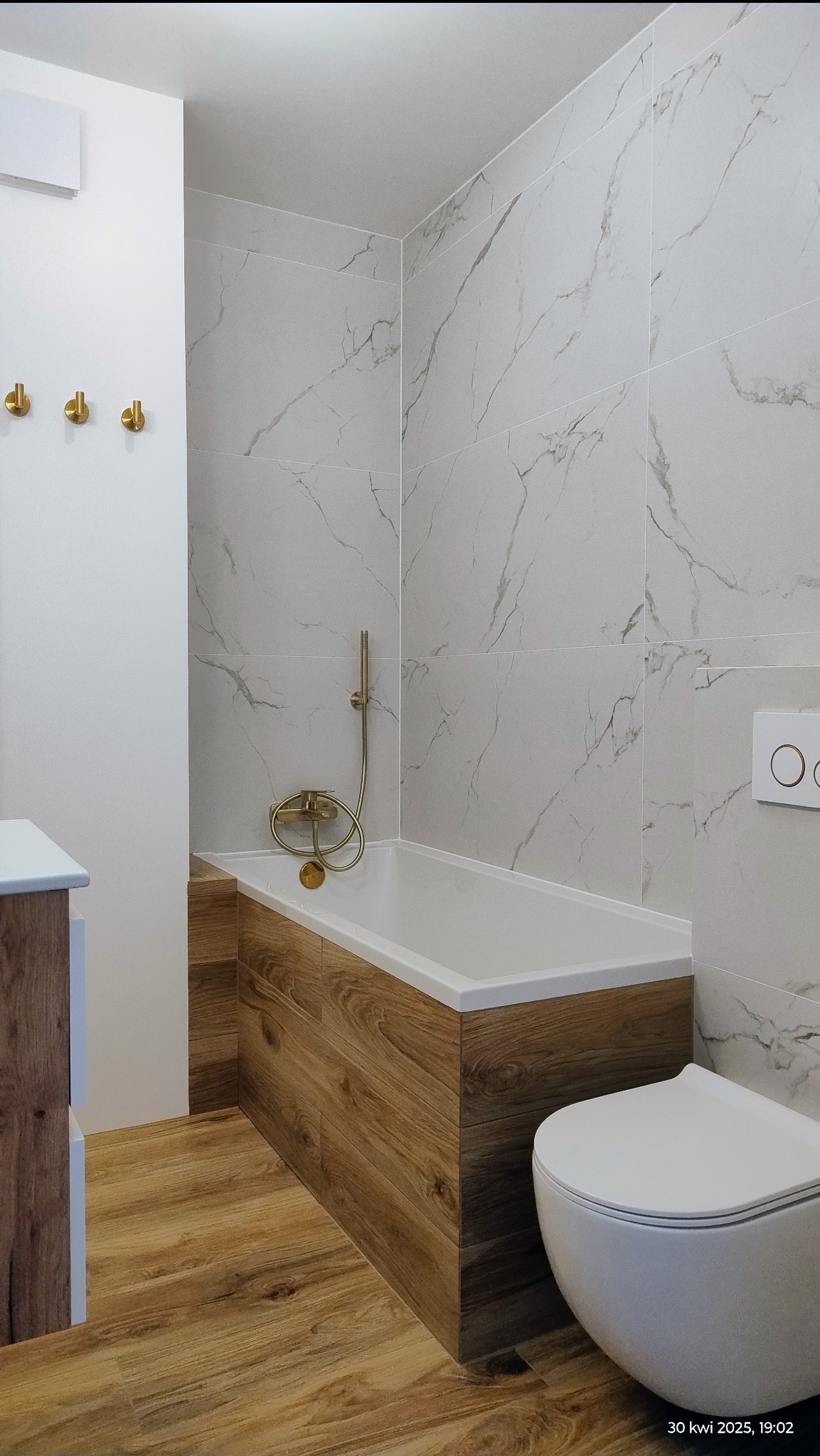 Nowoczesna łazienka z wanną obudowaną drewnopodobnymi panelami, marmurowe płytki na ścianach i złota armatura, tworzą elegancki i spójny wygląd.