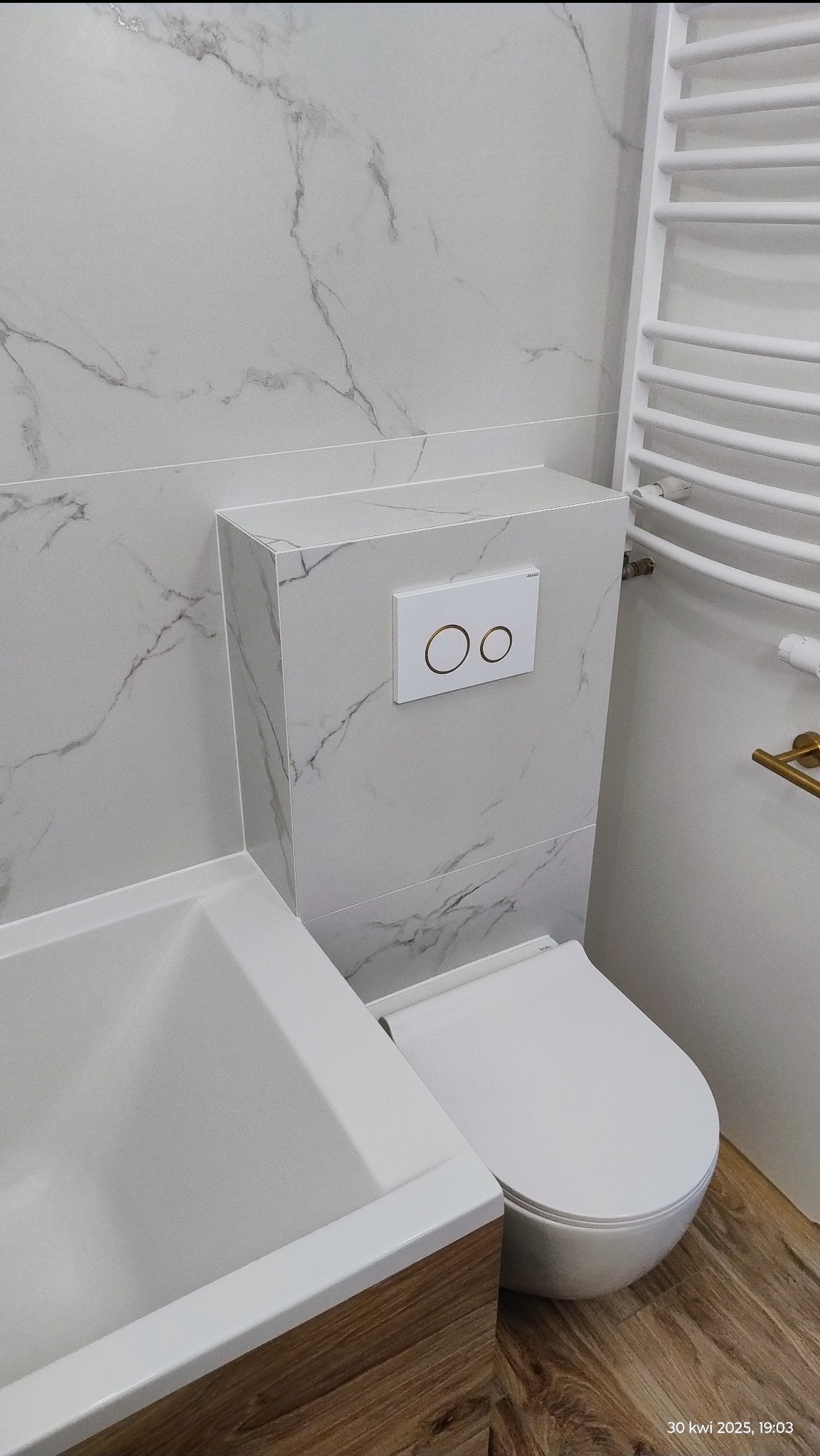 Nowoczesna łazienka w bieli i szarości z marmurowym wzorem na ścianach. Uwagę przykuwa wisząca toaleta z minimalistycznym przyciskiem spłukującym i drewniana szafka umywalkowa.