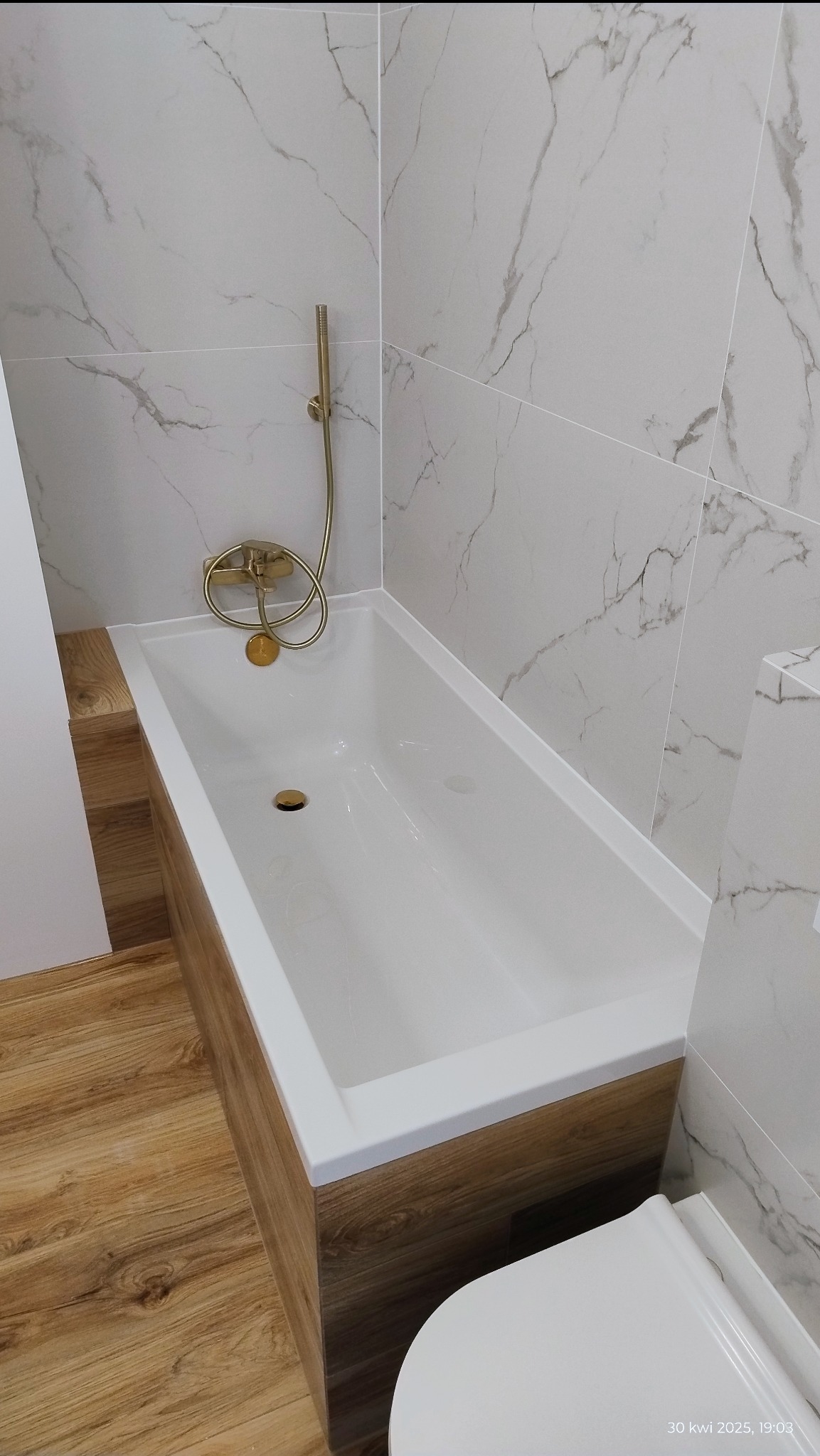 Biała wanna z obudową w odcieniach drewna, złota armatura, marmurowe płytki na ścianach i drewnopodobna podłoga tworzą elegancką aranżację łazienki.