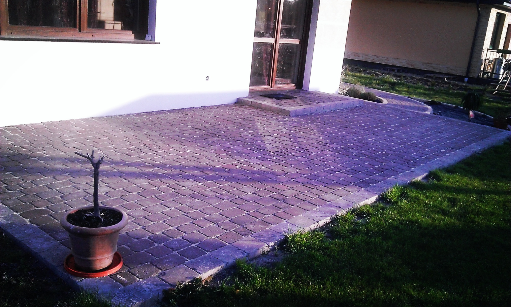 Ułożona z ciemnej kostki brukowej powierzchnia przed domem z donicą z uschniętą rośliną w lewym rogu.
