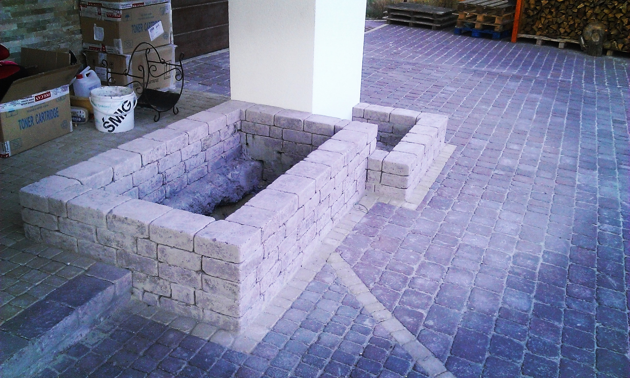 Budowa paleniska z kostki brukowej na patio, widoczne puste wnętrze i częściowo ułożona nawierzchnia z kostki, w tle palety z drewnem i kartony.