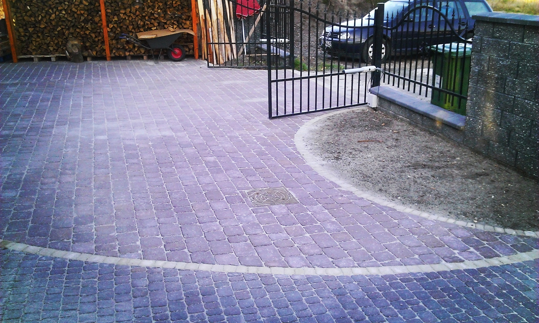 Ułożona kostka brukowa w odcieniach fioletu na podjeździe z otwartą, metalową bramą w tle, widoczny wózek i stos drewna.