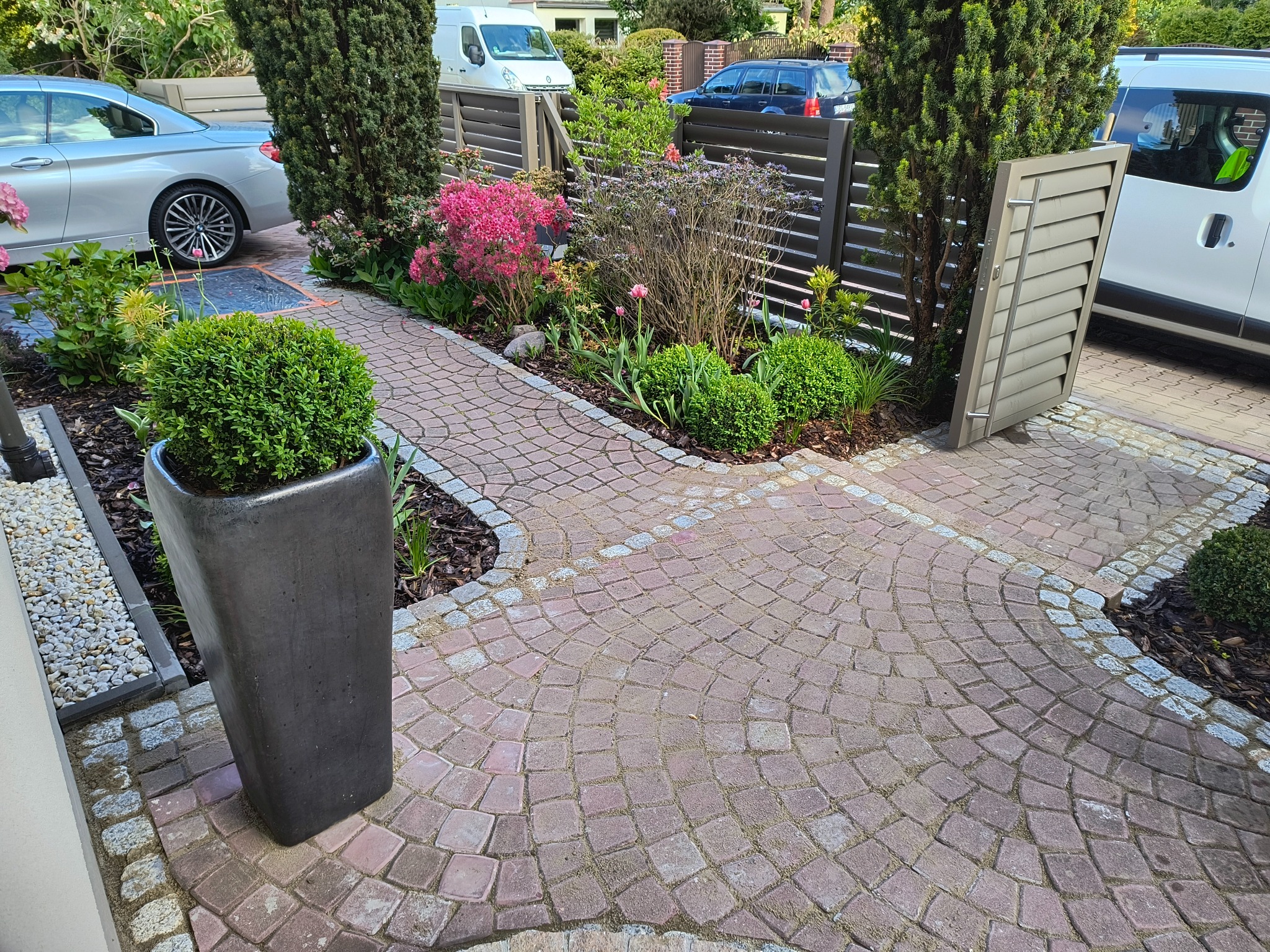 Kaskadowa ścieżka z kostki brukowej w odcieniach czerwieni, obrzeża z kostki granitowej, nowoczesne ogrodzenie panelowe i roślinność w zadbanym ogrodzie przydomowym.