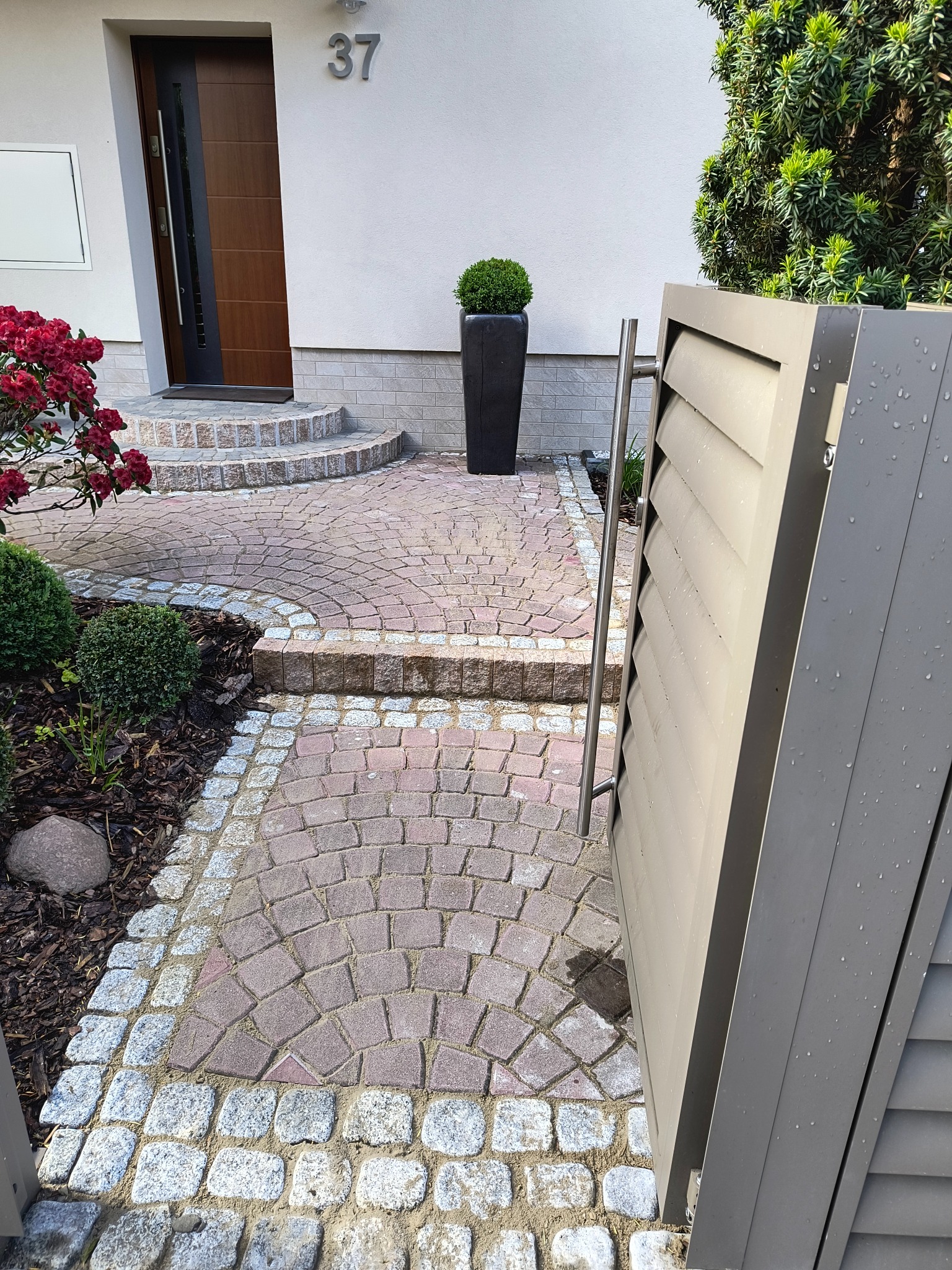 Ogród z kostki granitowej i brukowej prowadzący do domu z numerem 37, obok nowoczesne ogrodzenie lamelowe w kolorze szarym i roślinność. Wrocław, ul. Borówczana.