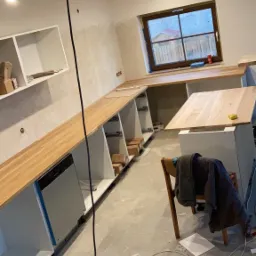 Montaż blatu drewnianego na szafkach kuchennych w surowym stanie, widoczne elementy instalacji elektrycznej i narzędzia na podłodze.