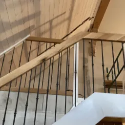 Wnętrze w trakcie budowy: drewniane schody z czarnymi, metalowymi balustradami, białe słupki, surowy drewniany strop, zabezpieczone folią elementy.